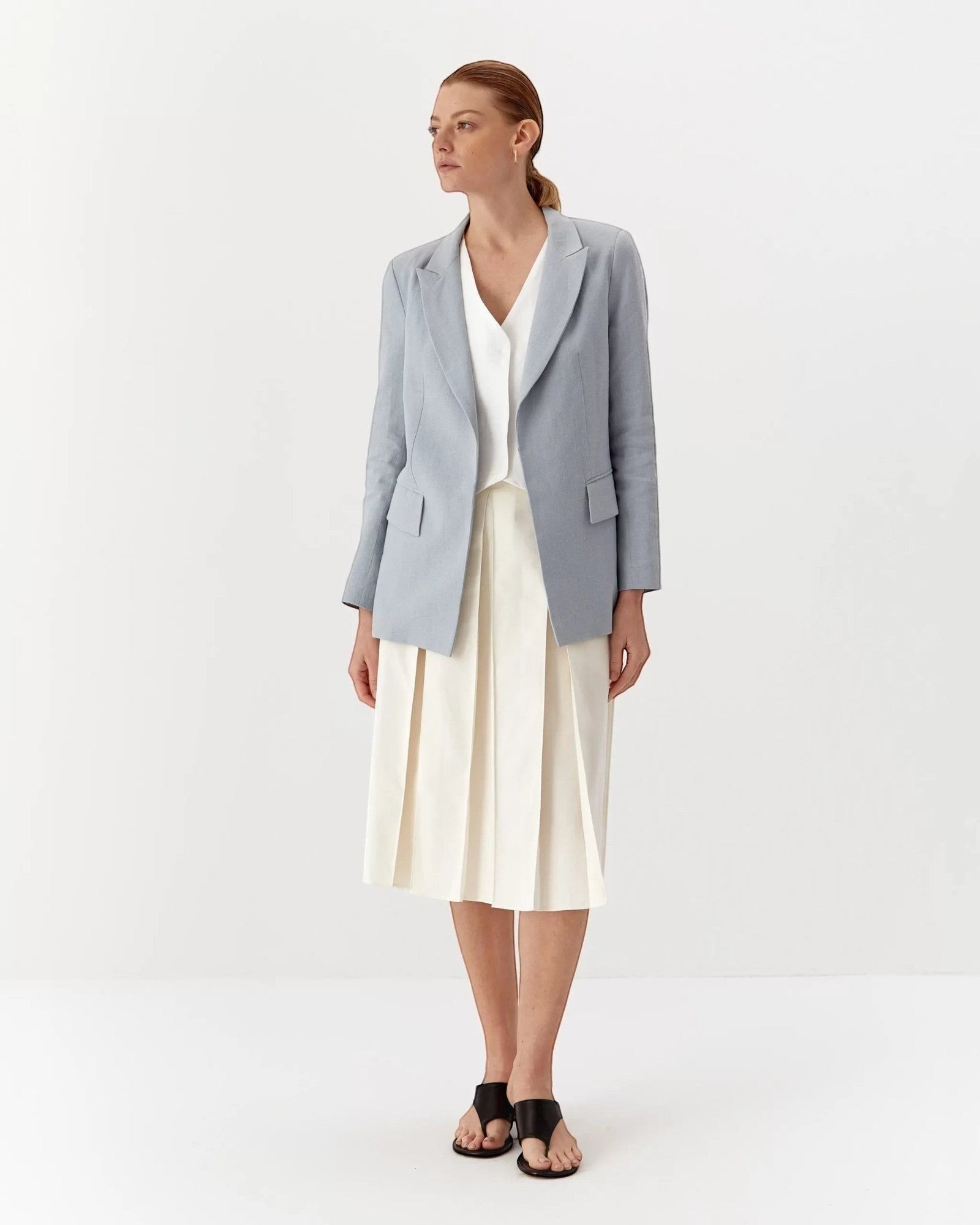 Ipanomi light blue linen blazer with lapel collar, flap pockets, and a tailored fit. Worn by a model with a white top and cream skirt.
