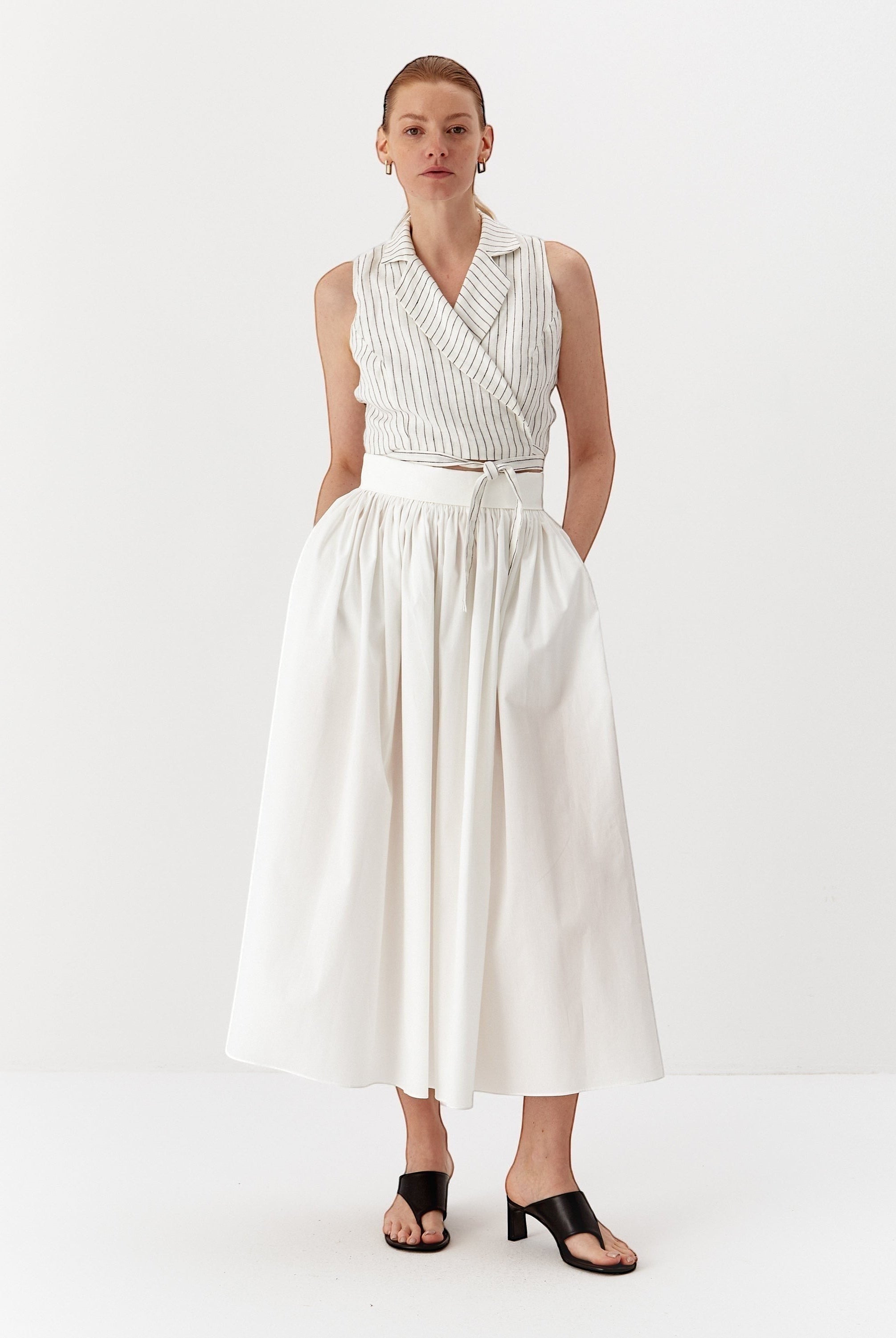Ipanomi white linen wrap vest with fine black pinstripes and a tie waist, paired with a white midi skirt.