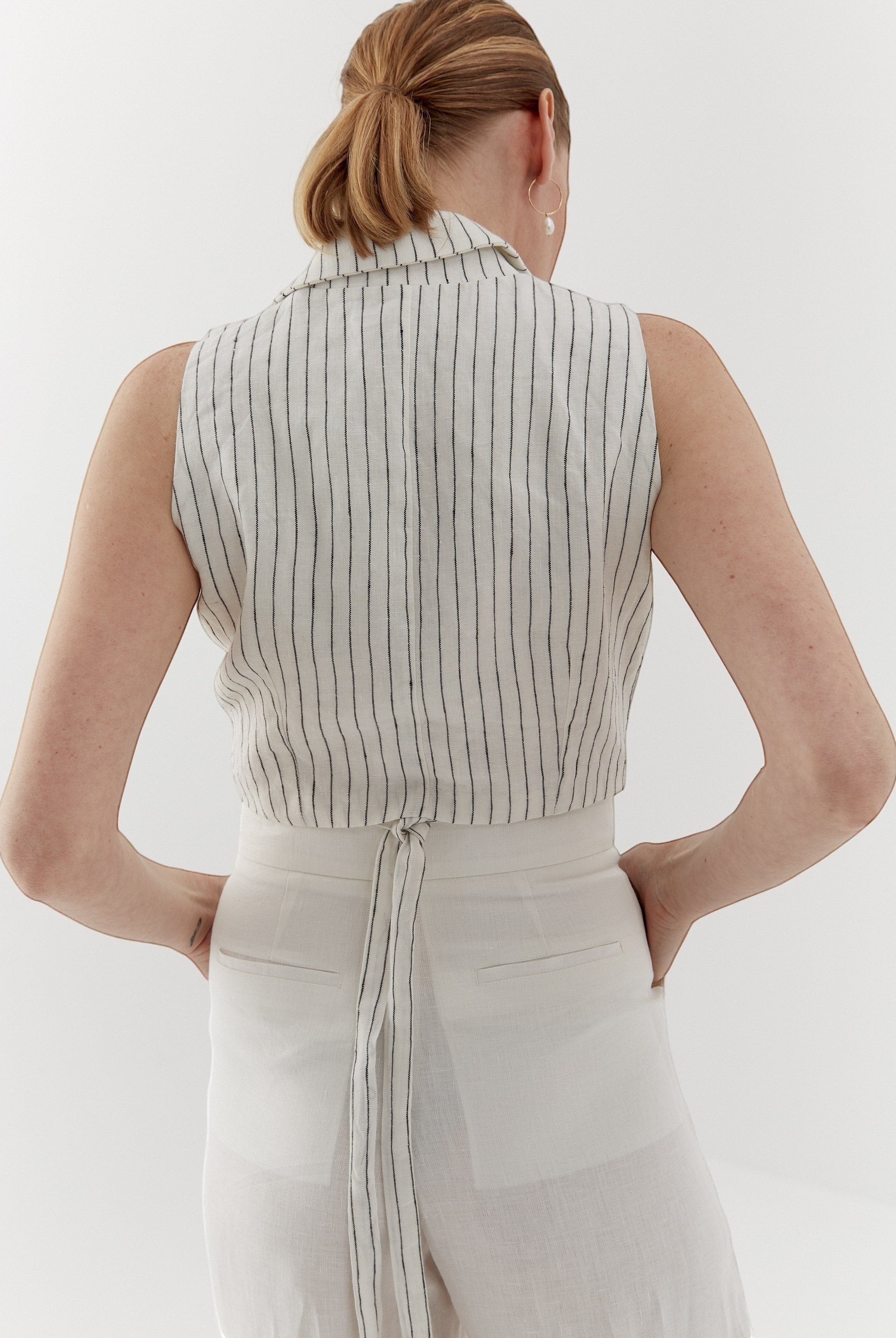 Ipanomi white linen vest with fine black pinstripes, featuring a wrap closure and adjustable tie back. Model wears size S.