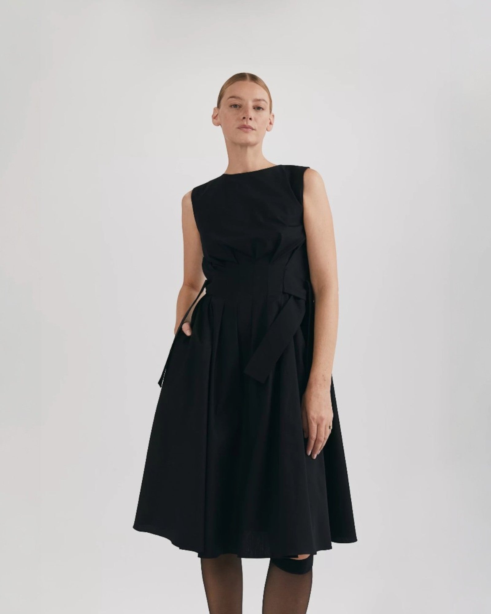 Ipanomi black organic cotton midi dress with pleats, adjustable tie, and hidden pockets. Sleeveless, round neck.