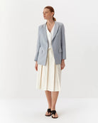 Ipanomi light blue linen blazer with lapel collar, flap pockets, and a tailored fit. Worn by a model with a white top and cream skirt.
