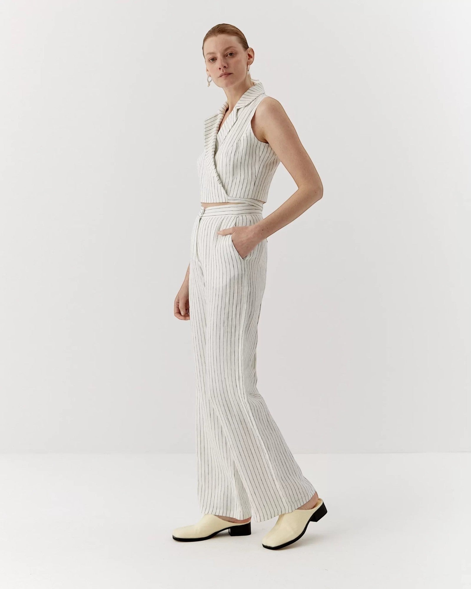 Ipanomi white linen trousers with black pinstripes, featuring a high waist, straight leg, and side pockets. Model wears size S.