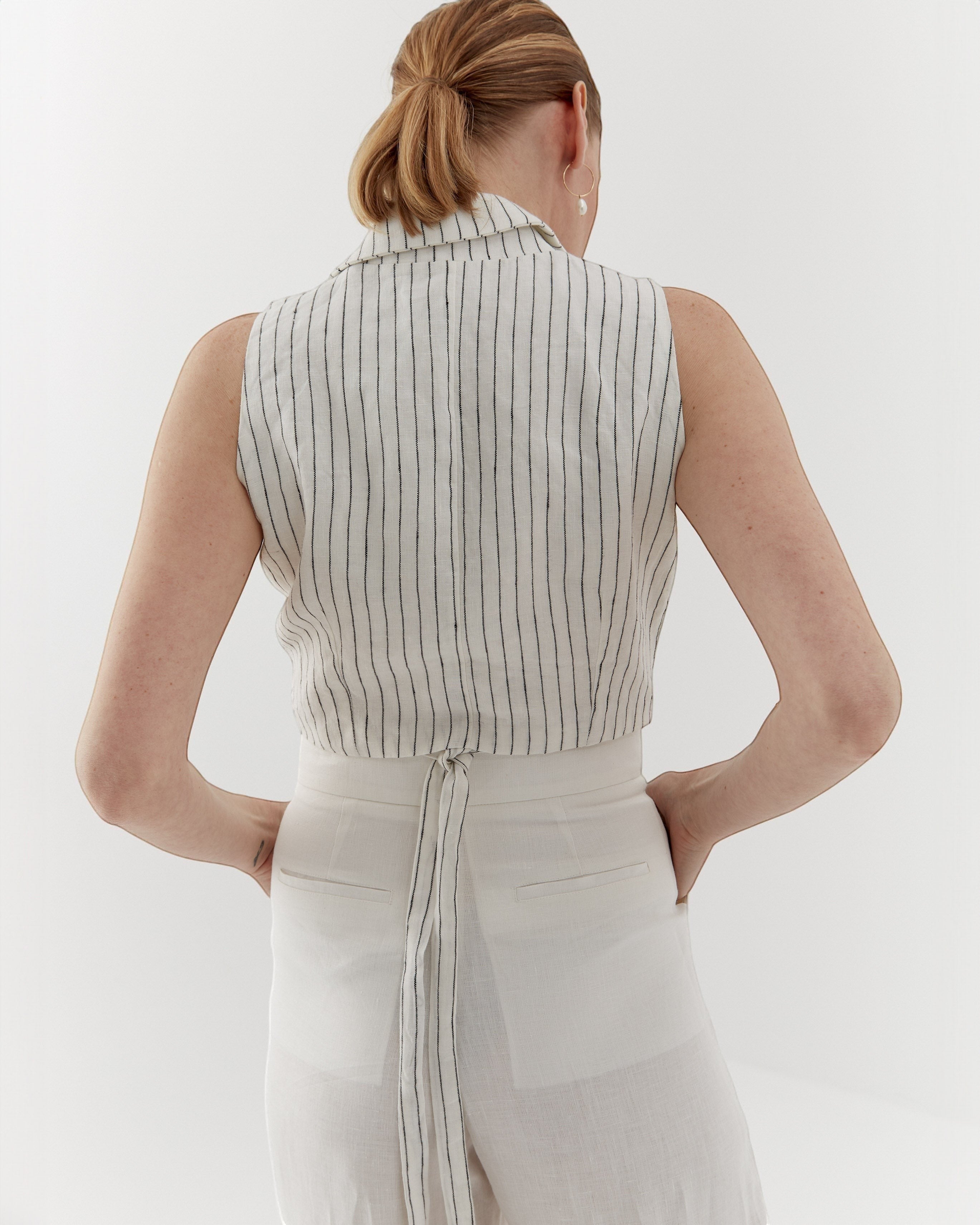 Ipanomi white linen vest with fine black pinstripes, featuring a wrap closure and adjustable tie back. Model wears size S.